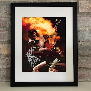 May include: Framed print featuring a rock musician playing a guitar, engulfed in fiery orange and yellow smoke. The image includes the band's logo and a signature. The musician is wearing a silver and black costume with silver boots.