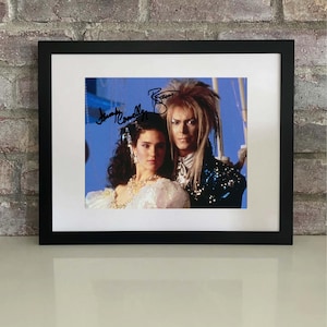 May include: Framed photograph featuring two people, one with long, spiky blonde hair and the other with dark hair and a white dress. The photograph is signed by both individuals. The frame is black, and the photograph is set against a white mat.