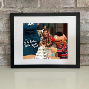 Puede incluir: Fotografía enmarcada con el Monstruo de las Galletas, Epi y un hombre cortando un pastel. El pastel está decorado con rayas de arcoíris y el número 30. El texto en el pastel dice "30th Anniversary". La fotografía está en un marco negro.