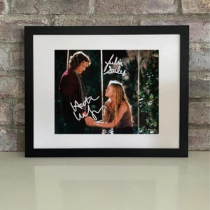 May include: Framed photograph featuring two people holding hands, with signatures in white ink. The photo is set against a brick wall. The frame is black, and the photo has a white border.