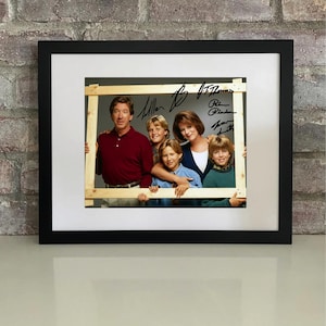 Puede incluir: Fotografía enmarcada con seis personas, incluyendo un hombre con camisa granate y una mujer con top azul. La foto está rodeada por un marco de madera e incluye firmas. El marco está sobre una pared de ladrillos.