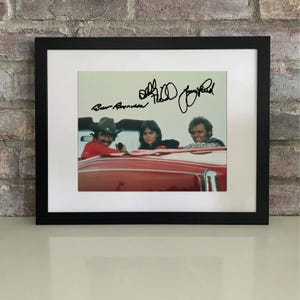 May include: Framed photograph featuring Burt Reynolds, Sally Field, and Jerry Reed in a red car. The image is signed by the actors. The black frame surrounds a white mat, enhancing the photograph's presentation. The image is set against a brick wall.