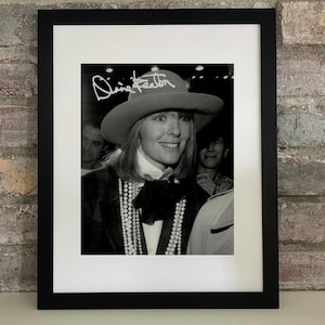 May include: Black and white framed photograph of Diane Keaton wearing a hat with her name on it. She is smiling and wearing a blazer, a bow tie, and a pearl necklace. The photograph is in a black frame.