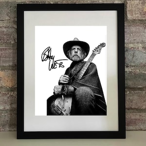 May include: Black and white framed print featuring a musician with a guitar. The musician is wearing a hat and poncho, holding a guitar. The print includes the signature "Bob Weir". The frame is black.