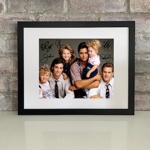 May include: Framed photograph featuring a cast photo from the television show "Full House." The image shows a group of people, with signatures in black ink. The frame is black, and the photograph is set against a white mat.