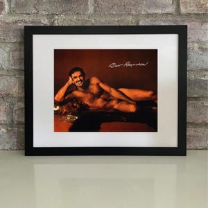 May include: Framed print of a man reclining on a fur throw, with a warm brown background. The man is smiling and holding a cigarette. The print is in a black frame. The image has the signature "Bur Reynolds".