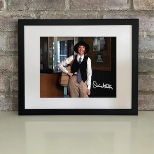 Puede incluir: Fotografía enmarcada de una persona con sombrero negro, chaleco y corbata, con camisa blanca y pantalones color caqui. La fotografía está firmada con tinta blanca. El marco es negro, sobre una pared de ladrillos.