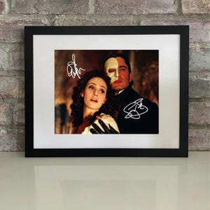 Könnte beinhalten: Gerahmter Druck mit einer Szene aus "Das Phantom der Oper". Das Bild zeigt zwei Schauspieler, einer davon mit einer Halbmaske, mit weißen Unterschriften. Der Druck ist in einem schwarzen Rahmen vor einer Backsteinmauer ausgestellt.