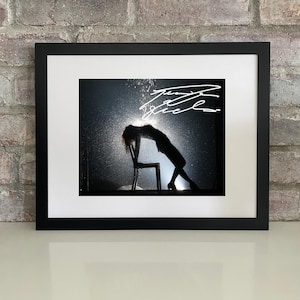 May include: Framed black and white photograph featuring a person leaning over a chair, with water droplets falling. The image has a signature in white. The frame is black, and the background is a brick wall.