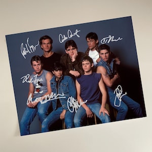 May include: A photograph featuring a group of eight men posing together. The image has several signatures in white ink. The men are wearing various outfits, including denim jackets, t-shirts, and tank tops. The background is a solid blue-gray color.