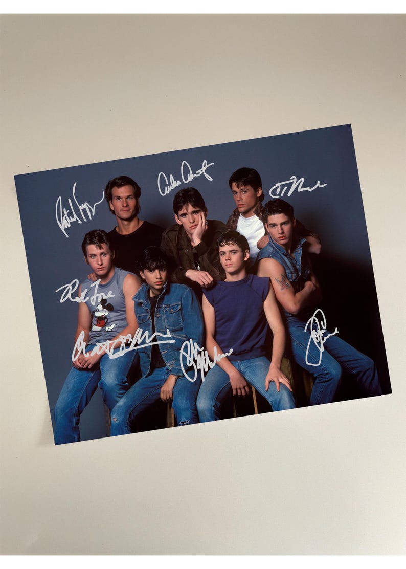 May include: A photograph featuring a group of eight men posing together. The image has several signatures in white ink. The men are wearing various outfits, including denim jackets, t-shirts, and tank tops. The background is a solid blue-gray color.
