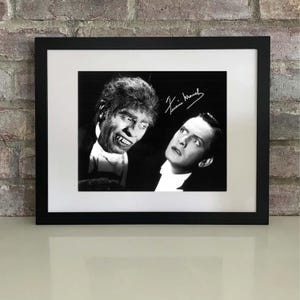 Puede incluir: Impresión enmarcada en blanco y negro con dos figuras. Una es un personaje parecido a un hombre lobo, y la otra es un hombre con traje. La impresión incluye una firma en la esquina superior derecha. El marco es negro y la impresión está sobre un passe-partout blanco.