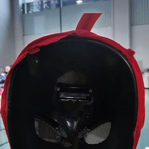 Puede incluir: Una m&aacute;scara negra con un borde de tela roja y una correa roja. La m&aacute;scara tiene agujeros para los ojos con un dise&ntilde;o de malla y una caracter&iacute;stica rectangular en la parte superior. La m&aacute;scara est&aacute; sujeta por una mano.