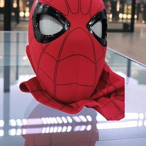 Puede incluir: Una m&aacute;scara roja de Spider-Man con detalles negros y cubiertas oculares de malla blanca. La m&aacute;scara tiene un patr&oacute;n de telara&ntilde;a y una cobertura para el cuello. La m&aacute;scara se exhibe sobre una superficie reflectante.