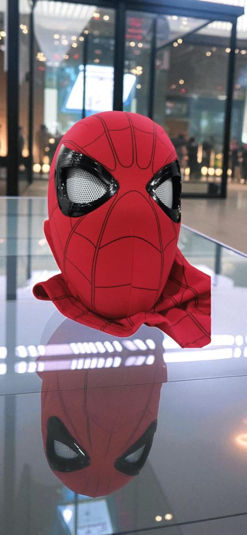 Puede incluir: Una m&aacute;scara roja de Spider-Man con detalles negros y cubiertas oculares de malla blanca. La m&aacute;scara tiene un patr&oacute;n de telara&ntilde;a y una cobertura para el cuello. La m&aacute;scara se exhibe sobre una superficie reflectante.