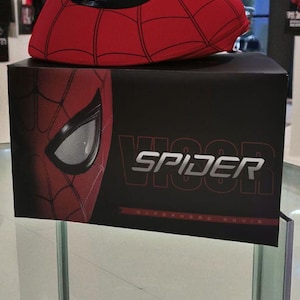 Puede incluir: Un sombrero de visera rojo de Spider-Man con una abertura para los ojos de malla negra se encuentra sobre una caja negra. La caja presenta la m&aacute;scara de Spider-Man y la palabra "SPIDER" en letras blancas.