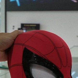 Puede incluir: Una m&aacute;scara roja de Spider-Man con telara&ntilde;as negras y grandes cubiertas oculares negras. Las cubiertas oculares tienen un material de malla blanca. La m&aacute;scara est&aacute; sujeta por una mano. El fondo est&aacute; borroso.