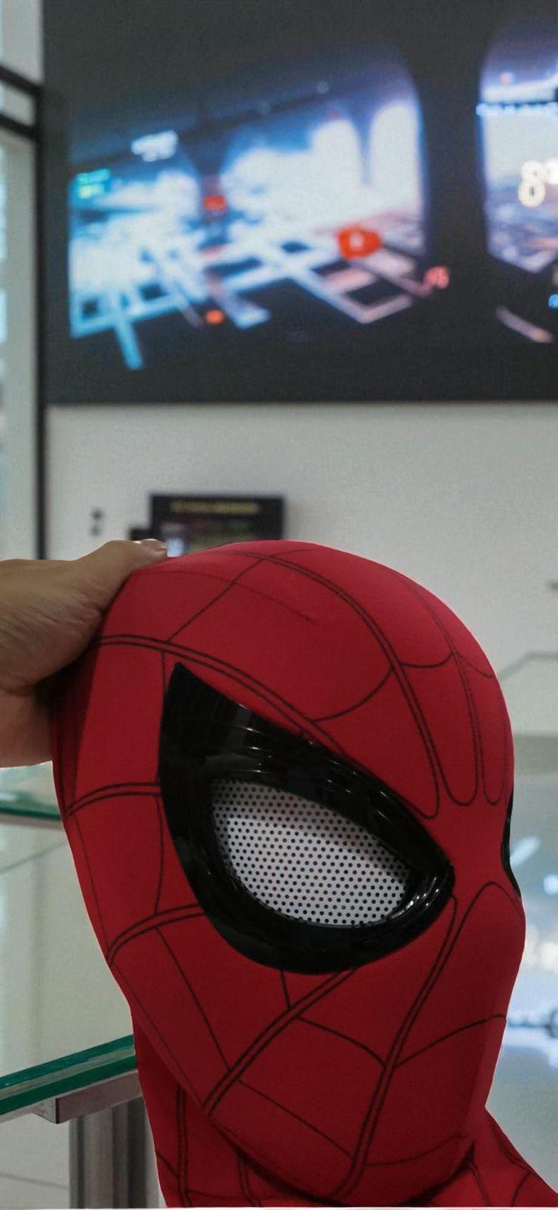Puede incluir: Una m&aacute;scara roja de Spider-Man con telara&ntilde;as negras y grandes cubiertas oculares negras. Las cubiertas oculares tienen un material de malla blanca. La m&aacute;scara est&aacute; sujeta por una mano. El fondo est&aacute; borroso.