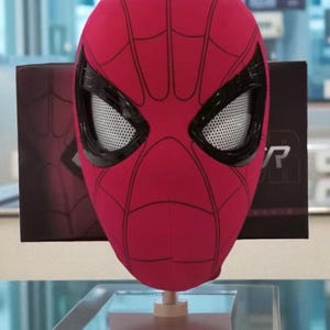 Puede incluir: Una m&aacute;scara roja de Spider-Man con detalles negros y un dise&ntilde;o de telara&ntilde;a. La m&aacute;scara se exhibe sobre un soporte, con una caja negra con el logotipo de Spider-Man en el fondo.