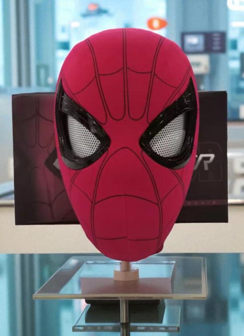 Puede incluir: Una m&aacute;scara roja de Spider-Man con detalles negros y un dise&ntilde;o de telara&ntilde;a. La m&aacute;scara se exhibe sobre un soporte, con una caja negra con el logotipo de Spider-Man en el fondo.
