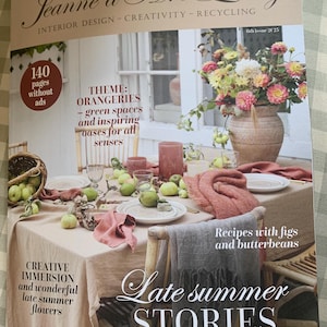 May include: A magazine cover with the title "Jeanne d'Arc Living" featuring interior design. The cover showcases a table setting with apples, flowers, and a pink tablecloth. Text includes "Late Summer Stories" and "Recipes with figs and butterbeans."