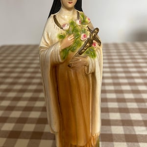 May include: A religious figurine of a saint wearing a black veil and a cream-colored habit, holding a brown crucifix and a bouquet of pink roses with green leaves. The figure stands on a small green base with painted pink floral accents.