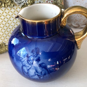 May include: A blue and gold ceramic pitcher with a white interior and a floral design. The pitcher has a round body and a curved handle.