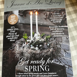 May include: A magazine cover titled "Jeanne d'Arc Living" with a spring theme. It features a nest-like arrangement with eggs, greenery, and two lit candles. The cover includes text about interior design, creativity, and recycling, with the words "Get ready for SPRING".
