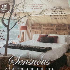 May include: A magazine cover with the text "Jeanne d'Arc Living" and "Sensuous SUMMER." The image features a bed with a carved wooden headboard, white bedding, and decorative pillows. A tree trunk serves as a side table, and a lamp with a red shade is visible.