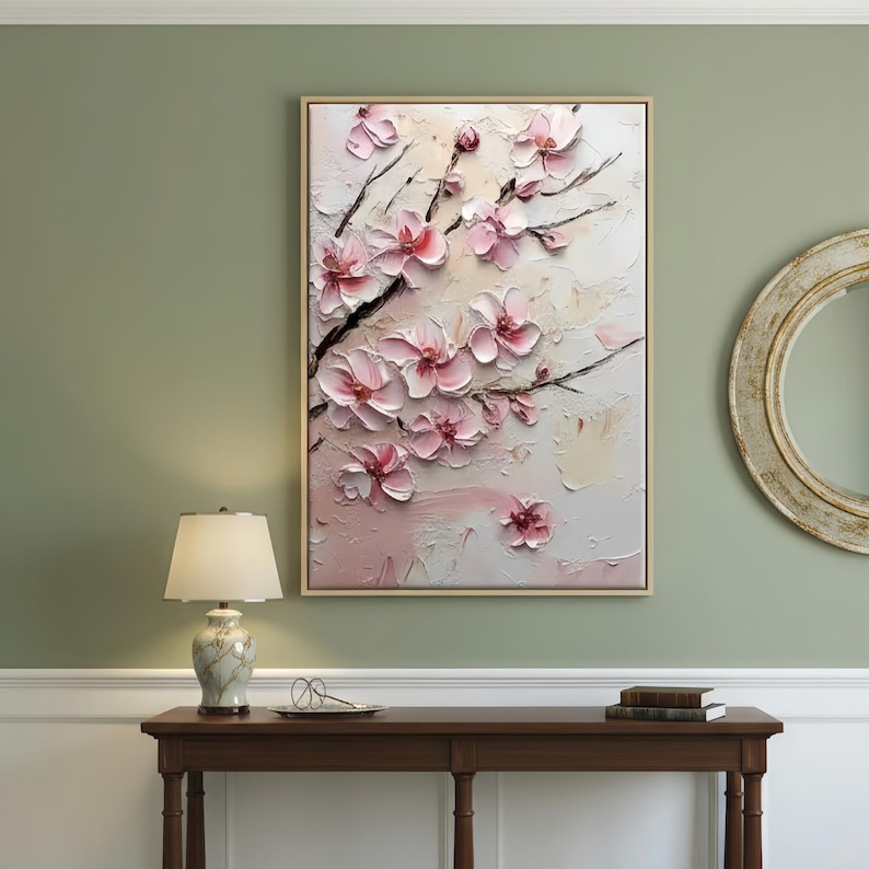 May include: A framed floral painting featuring pink blossoms on dark branches against a textured, off-white background. The artwork is displayed above a wooden table with a lamp and decorative items.