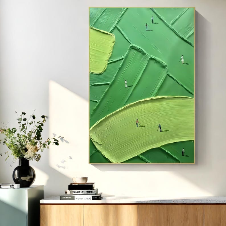 May include: Abstract painting of a golf course in shades of green, featuring textured brushstrokes and miniature golfer figures. The artwork is framed in a gold frame and hangs on a white wall.