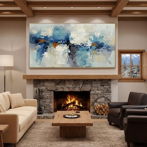 May include: Abstract painting in a gold frame, with blue, white, and orange brushstrokes. It hangs above a stone fireplace with a burning fire, creating a cosy living room setting. The room includes a beige sofa, wooden coffee table, and leather armchairs. The painting is approximately 150cm x 75cm.