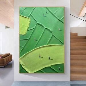 May include: Abstract oil painting on canvas featuring a golf course scene. The artwork uses shades of green with textured brushstrokes to depict the landscape. Miniature figures of golfers are scattered across the course, framed in a gold border.