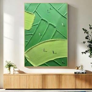 May include: Abstract green textured painting with a gold frame. The artwork features a layered design with varying shades of green and tiny figures scattered across the surface, suggesting a landscape or field. The painting is displayed above a wooden cabinet.