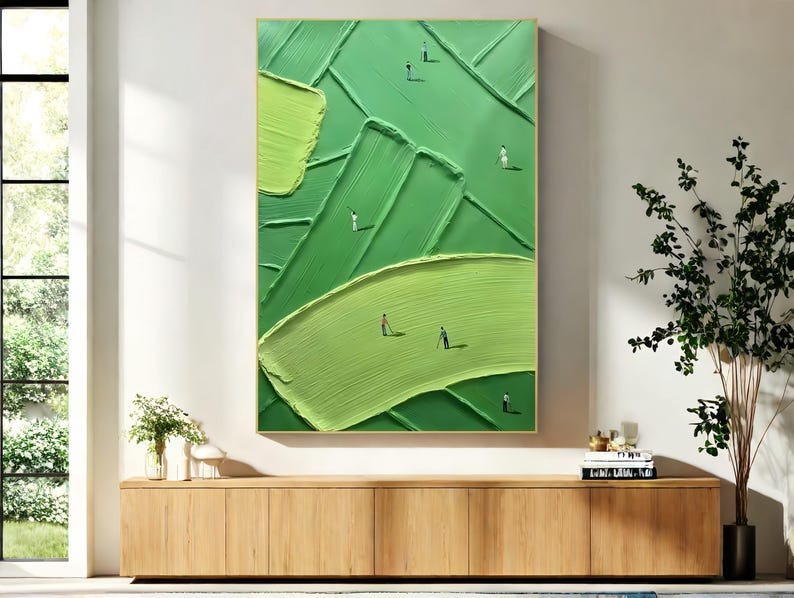 May include: Abstract green textured painting with a gold frame. The artwork features a layered design with varying shades of green and tiny figures scattered across the surface, suggesting a landscape or field. The painting is displayed above a wooden cabinet.