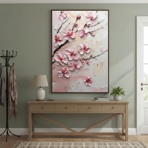 May include: A framed floral painting featuring pink cherry blossoms on a textured white and cream background. The artwork is displayed above a wooden console table with a lamp and decorative items. The room has a neutral color palette.