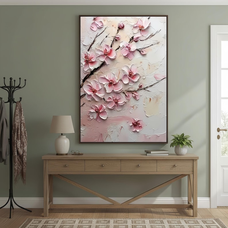 May include: A framed floral painting featuring pink cherry blossoms on a textured white and cream background. The artwork is displayed above a wooden console table with a lamp and decorative items. The room has a neutral color palette.