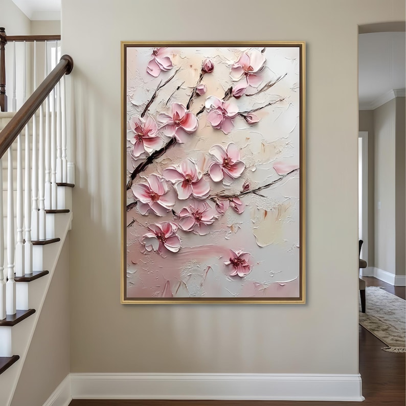 May include: A framed painting of pink blossom flowers on dark branches, set against a textured, off-white backdrop. The artwork is in a gold frame and hangs on a beige wall, offering a nature-inspired decorative element.