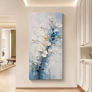 Puede incluir: Pintura vertical sobre lienzo que presenta un arreglo floral con flores blancas y azules. Las flores tienen una apariencia texturizada y en relieve sobre un fondo azul claro y blanco. La obra de arte se exhibe en una pared blanca.