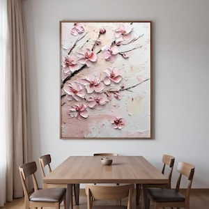 May include: A framed floral painting featuring textured, three-dimensional pink blossoms on dark branches. The artwork has a light pink and white background with visible brushstrokes. The painting is displayed above a wooden dining table.