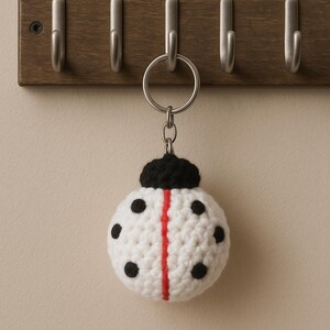 May include: A handmade white crochet ladybug keychain with black spots and a red stripe down the center. The keychain is attached to a silver metal ring and hangs from a wooden wall-mounted hook rack.