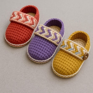May include: Three colorful crochet baby shoes in red, purple, and yellow. Each shoe has a white sole and a strap with a chevron pattern. The yellow shoe has a wooden button detail.