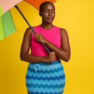 May include: A woman wearing a pink tank top and a blue crochet skirt, holding a rainbow umbrella against a yellow background. The skirt features a wavy pattern in shades of blue. The umbrella has a black handle.