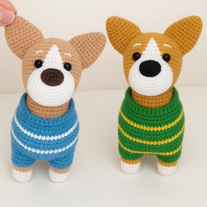 May include: Two handmade crochet dog figurines. One dog is tan with a blue sweater with white stripes. The other dog is tan with a green sweater with yellow stripes. Both have white faces, black eyes, and brown noses.