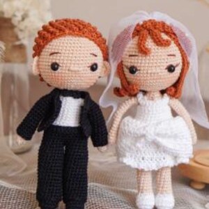 Puede incluir: Muñecos de novia y novio hechos a ganchillo. El novio lleva un traje negro con camisa blanca. La novia lleva un vestido blanco y velo. Ambos muñecos tienen pelo rojo y se dan la mano. Los muñecos miden unos 15 cm de alto.