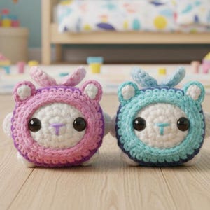 May include: Two handmade crochet toys, one pink and one blue, each with a bear-like hood and large black eyes. The toys are white with colorful accents and small, round ears. They are sitting on a wooden surface.