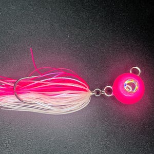 May include: A pink and white fishing jig with a hook and a chain connecting to a pink, round head. The jig has a colorful eye and pink and white skirt. The background is black.