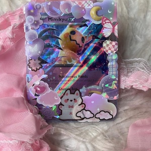Pode incluir: Um cartão Pokémon Mimikyu ex decorado com um personagem amarelo e preto, rodeado por enfeites rosa e roxos. O cartão apresenta o texto "Mischievous Hands" e "Ghost Trip". Os elementos decorativos incluem nuvens, corações e borboletas.