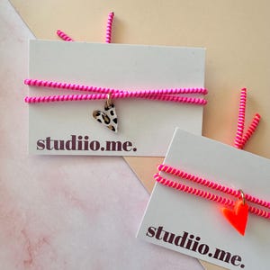 Könnte beinhalten: Zwei Armbänder mit leuchtend pinkfarbenen Spiralkordeln auf weißen Präsentationskarten mit dem Text studiio.me. Ein Armband hat einen herzförmigen Anhänger mit schwarz-beigem Leopardenmuster, das andere einen einfarbigen neonorangen Herzanhänger.