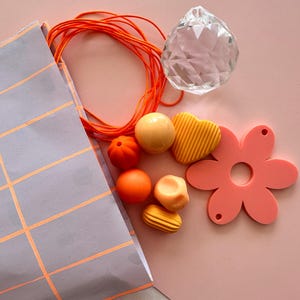 Könnte beinhalten: Eine Sammlung von Bastelmaterialien auf rosa Hintergrund. Enthält eine graue Tasche mit orangefarbenen Gitterlinien, orangefarbenes Garn, einen klaren Kristallkugel und verschiedene orange und gelbe Perlen und Formen, darunter eine Blume.